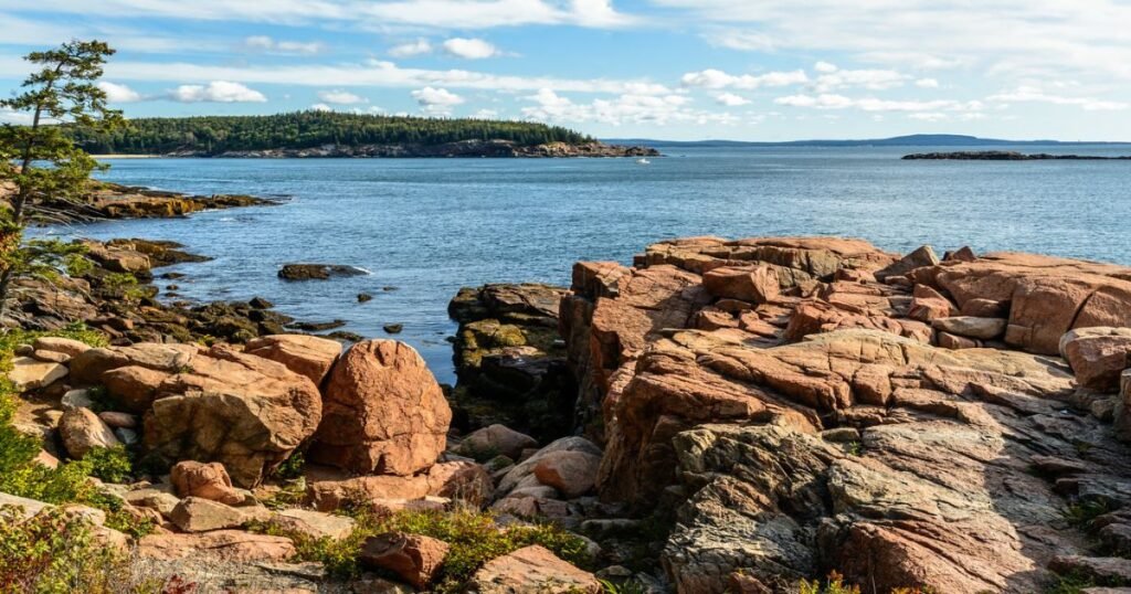 A view from Cadillac Mountain overlooking Acadia's rugged coastline at sunrise with soft clouds above.