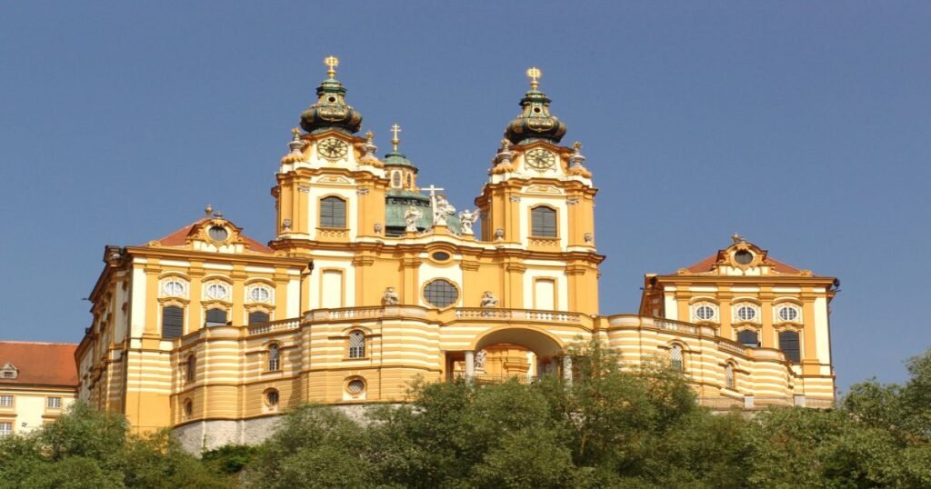 The golden façade of Melk Abbey standing proudly above the Danube River, surrounded by lush greenery.