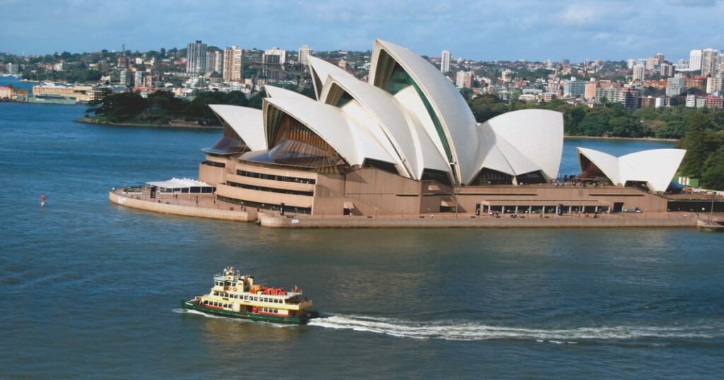 The iconic white sails of the Sydney Opera House stand out against a clear blue sky with the harbor in the foreground.