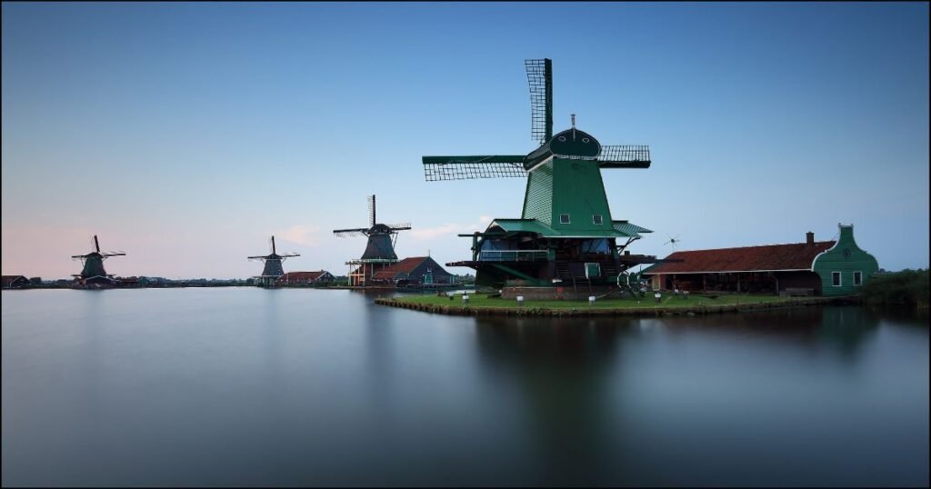  Traditional windmills standing along the riverbank in Zaanse Schans, surrounded by green fields and blue skies.