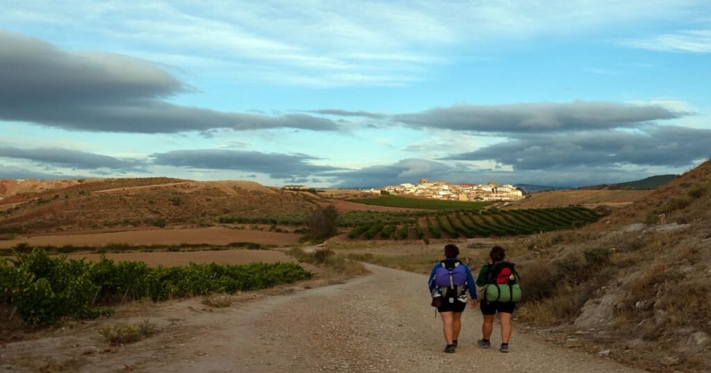  Pilgrims hiking along the scenic Camino de Santiago trail with rolling hills and a village in the distance.