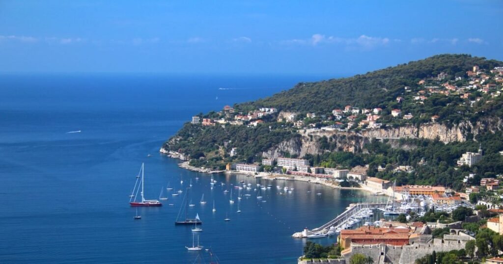 A view of the French Riviera’s sparkling blue waters, framed by palm trees and colorful buildings.