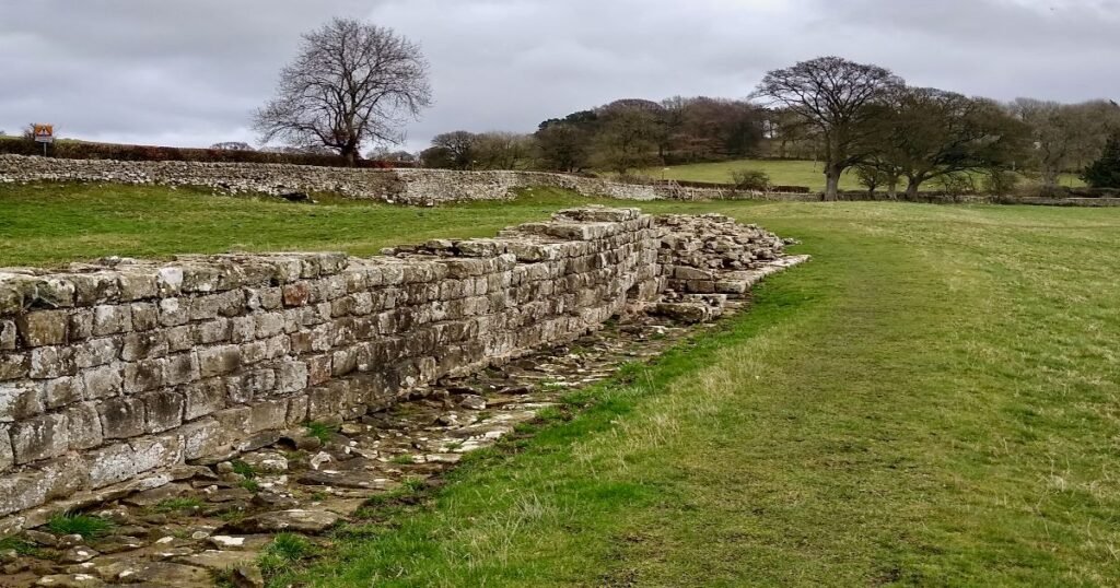 “Hadrian’s Wall cutting across the rolling hills of Northumberland, with ruins of ancient forts visible along the route.”