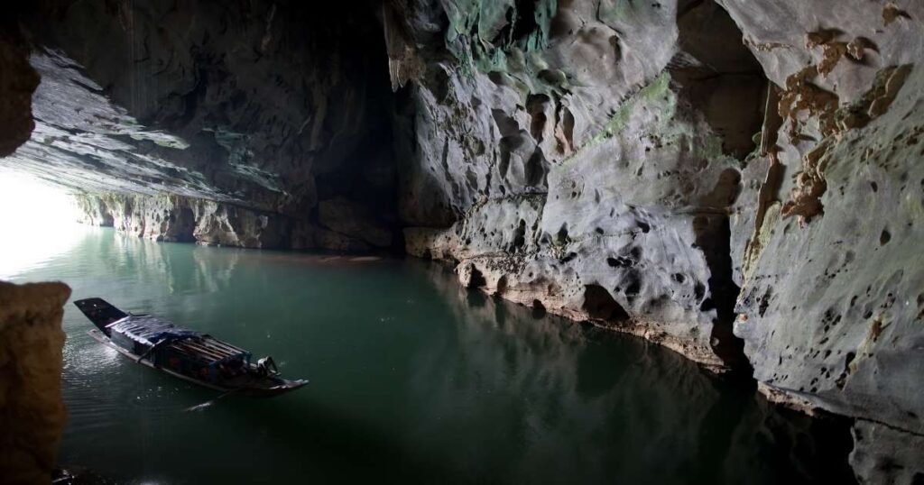 "Entrance to Phong Nha Cave, surrounded by lush greenery and a clear blue river in Phong Nha-Ke Bang National Park, Vietnam."