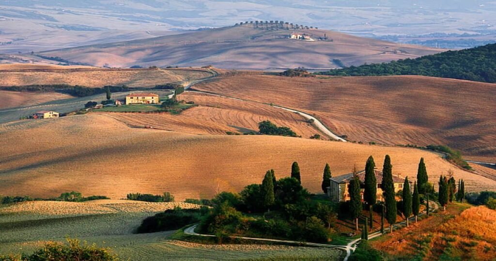 “Tuscany’s rolling hills with vineyards, olive groves, and cypress trees under a bright blue sky.”