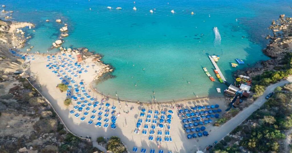 “Clear turquoise waters and golden sandy beach at Konnos Bay with rocky cliffs.”